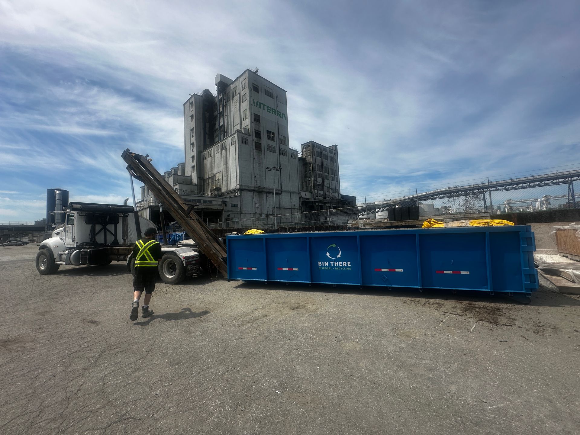 Worker near a blue conveyor and industrial silos on a gravel site under a cloudy sky