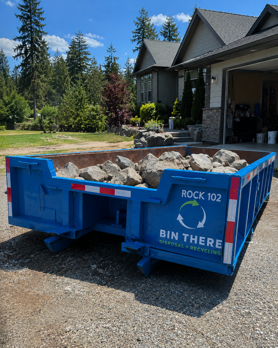 Blue dumpster filled with rocks on a gravel driveway beside a house and trees