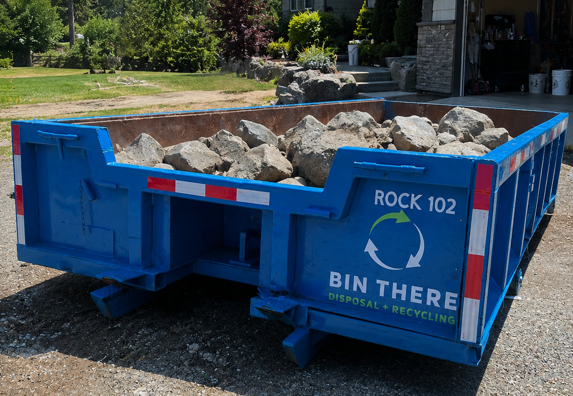 Blue dump trailer filled with rocks in a driveway beside a house under clear sky