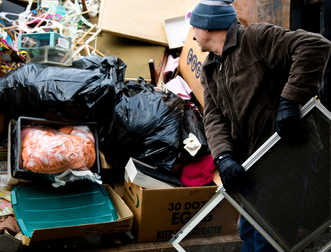 Person in winter clothes sorting through piled trash bags and boxes outdoors