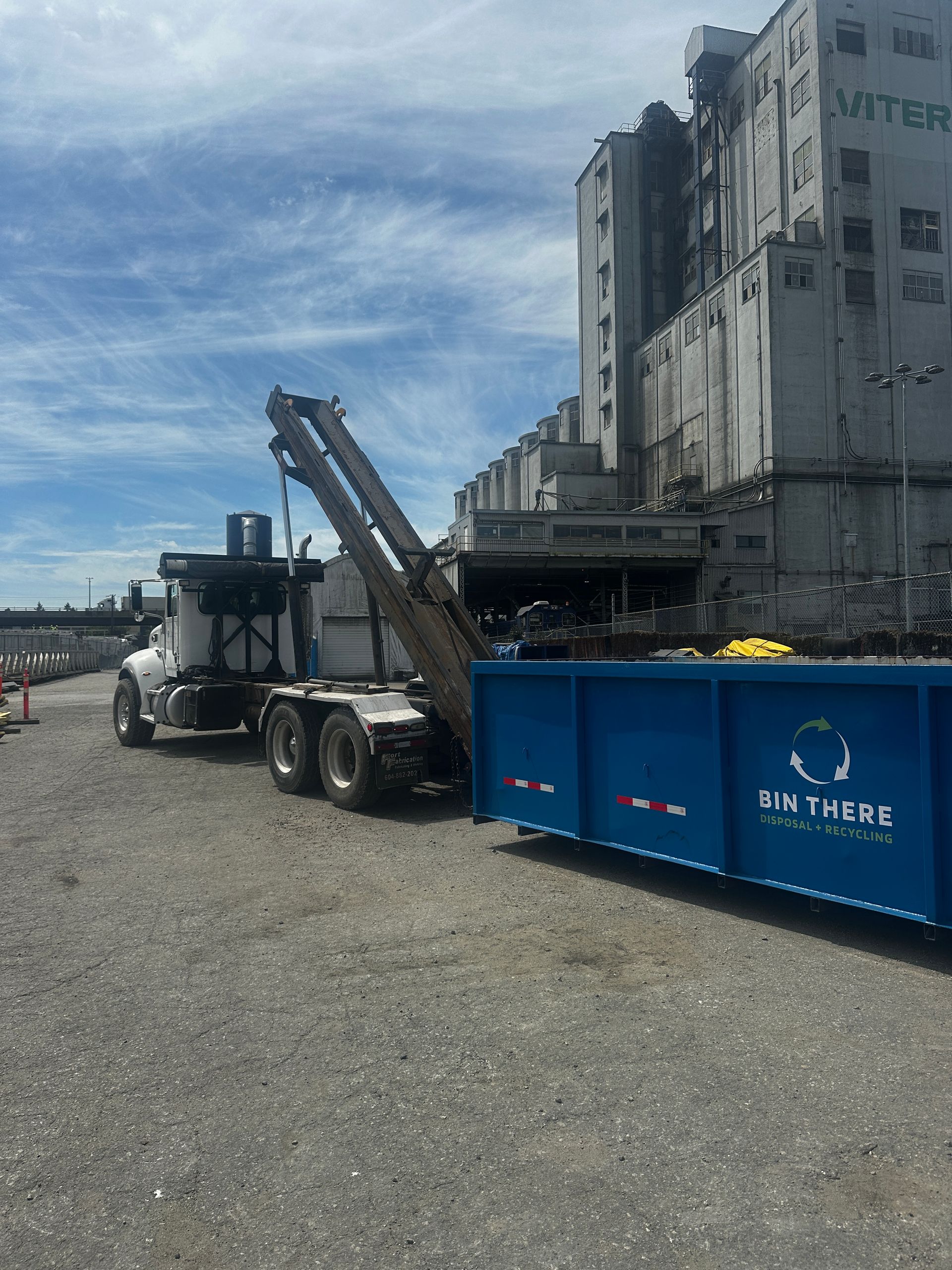 Construction truck beside blue dumpster near industrial building under cloudy sky
