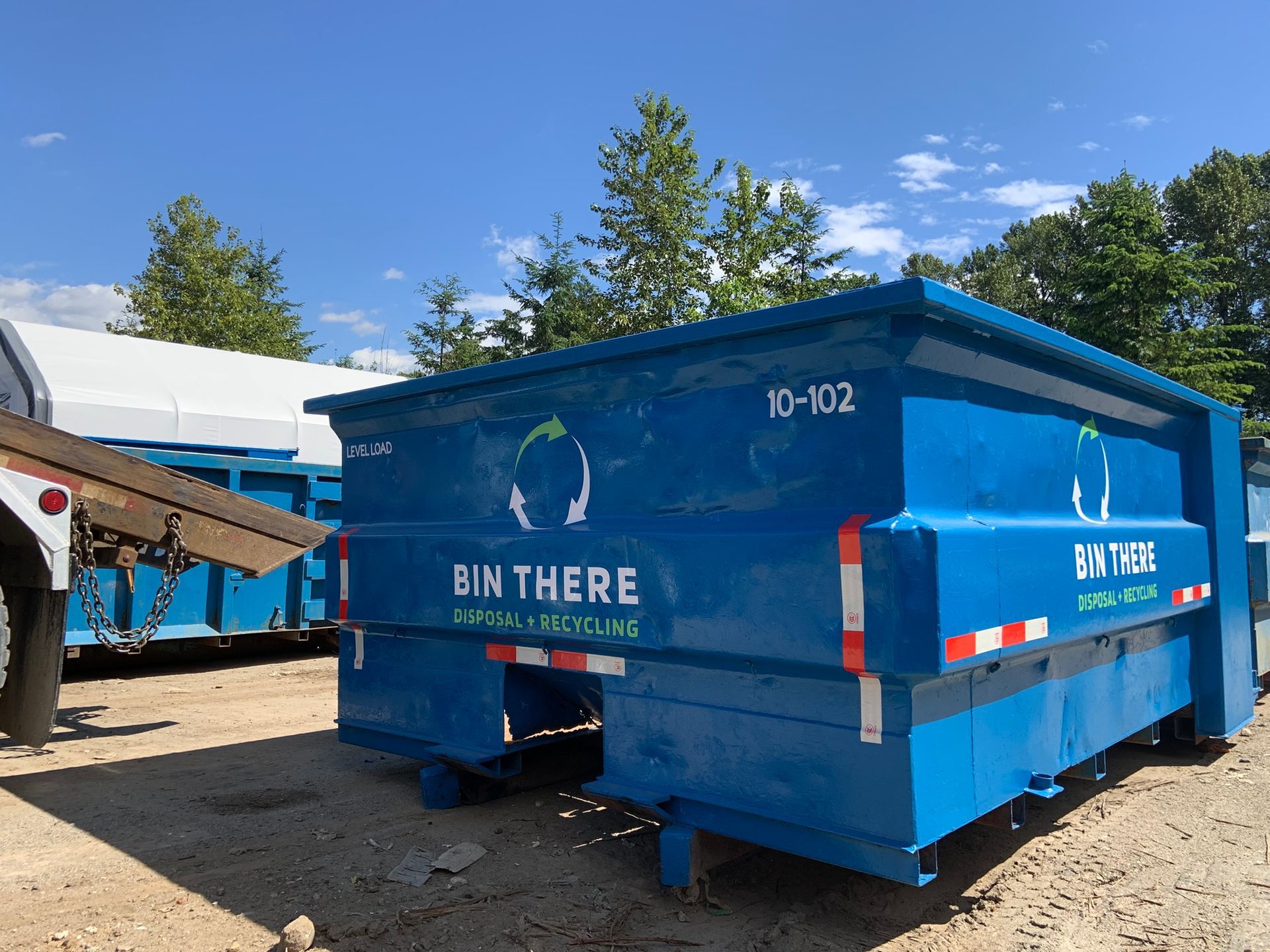 Blue dumpster in a sunny yard with “BIN THERE” lettering and a truck nearby
