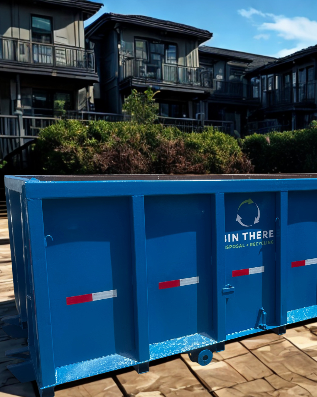 Blue construction dumpster in front of dark apartment buildings on a sunny day