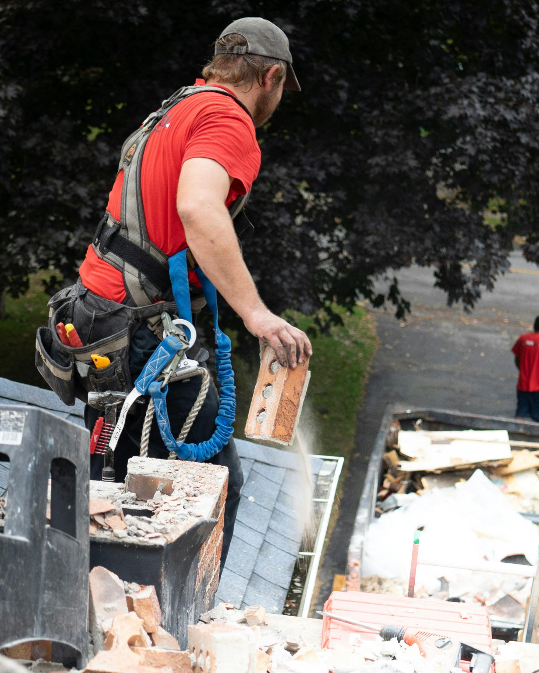 Worker in a red shirt and safety harness standing on a rooftop construction site carrying a brick block