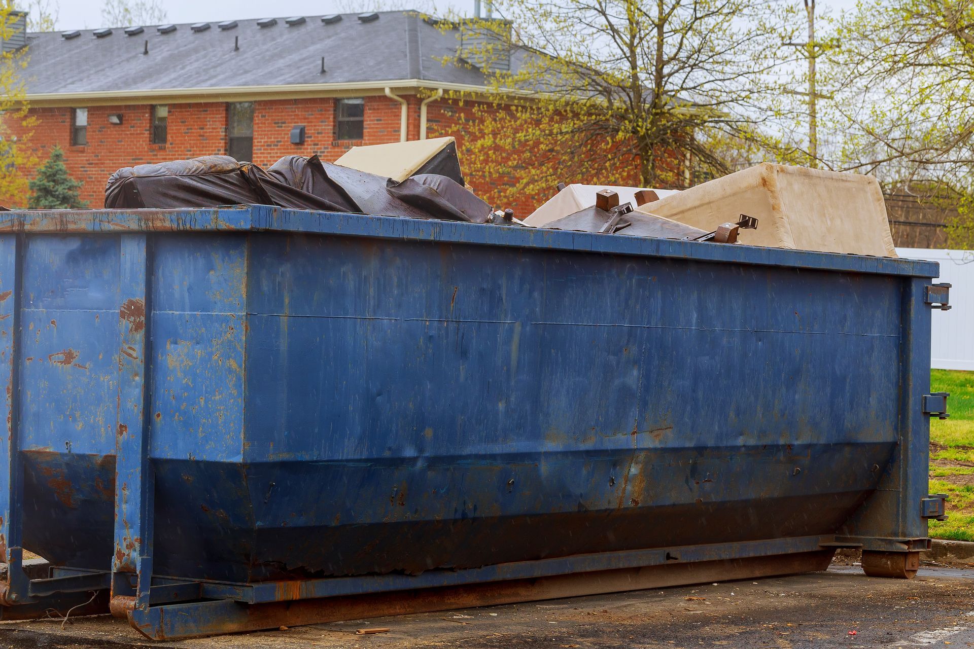 A large blue dumpster filled with trash is parked in front of a brick house.