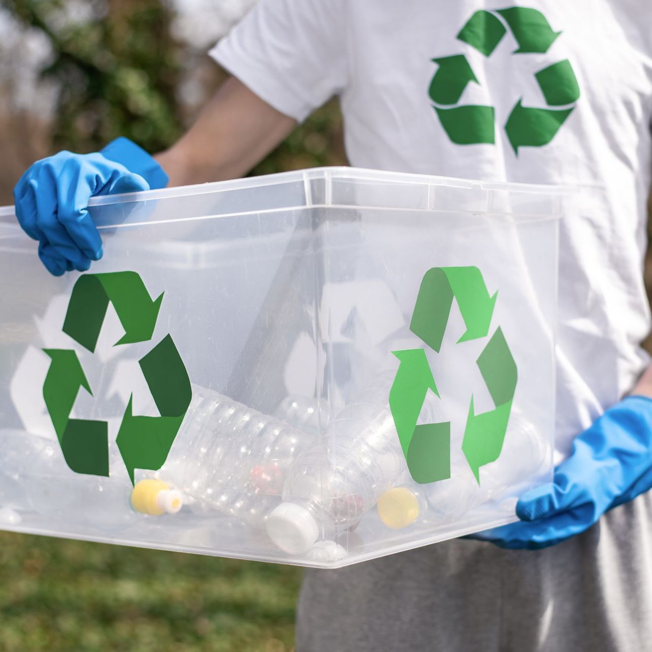 A person wearing blue gloves is holding a plastic container with recycling symbols on it