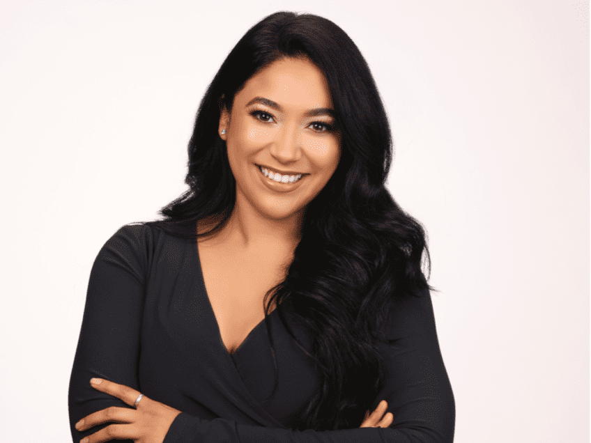 A woman in a black dress is smiling with her arms crossed.