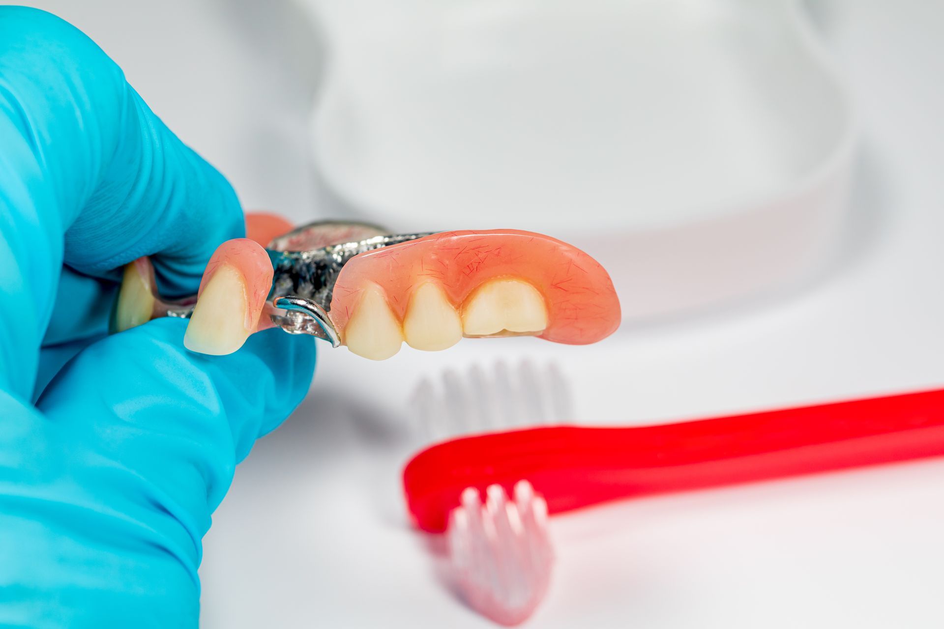 Gloved hand holding a partial denture beside a red toothbrush on a white surface Gloved hand holding a partial denture beside a red toothbrush on a white surface