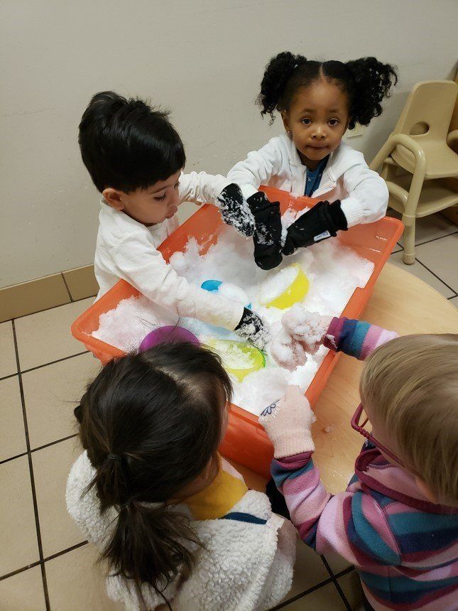 Children in white smocks mix colorful sensory foam at a table in a classroom. Children in white smocks mix colorful sensory foam at a table in a classroom.