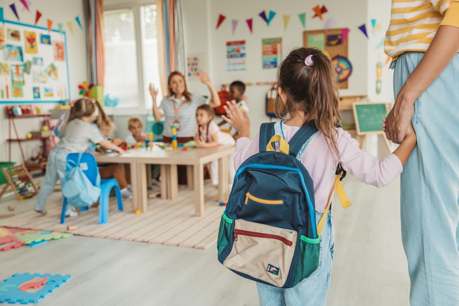 Child with backpack holding an adult’s hand in a bright classroom with children at tables and colorful bunting.
