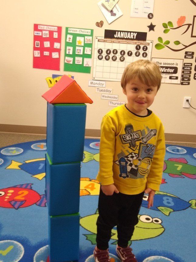 Child standing beside a tall blue block tower in a colorful classroom, smiling at the camera Child standing beside a tall blue block tower in a colorful classroom, smiling at the camera