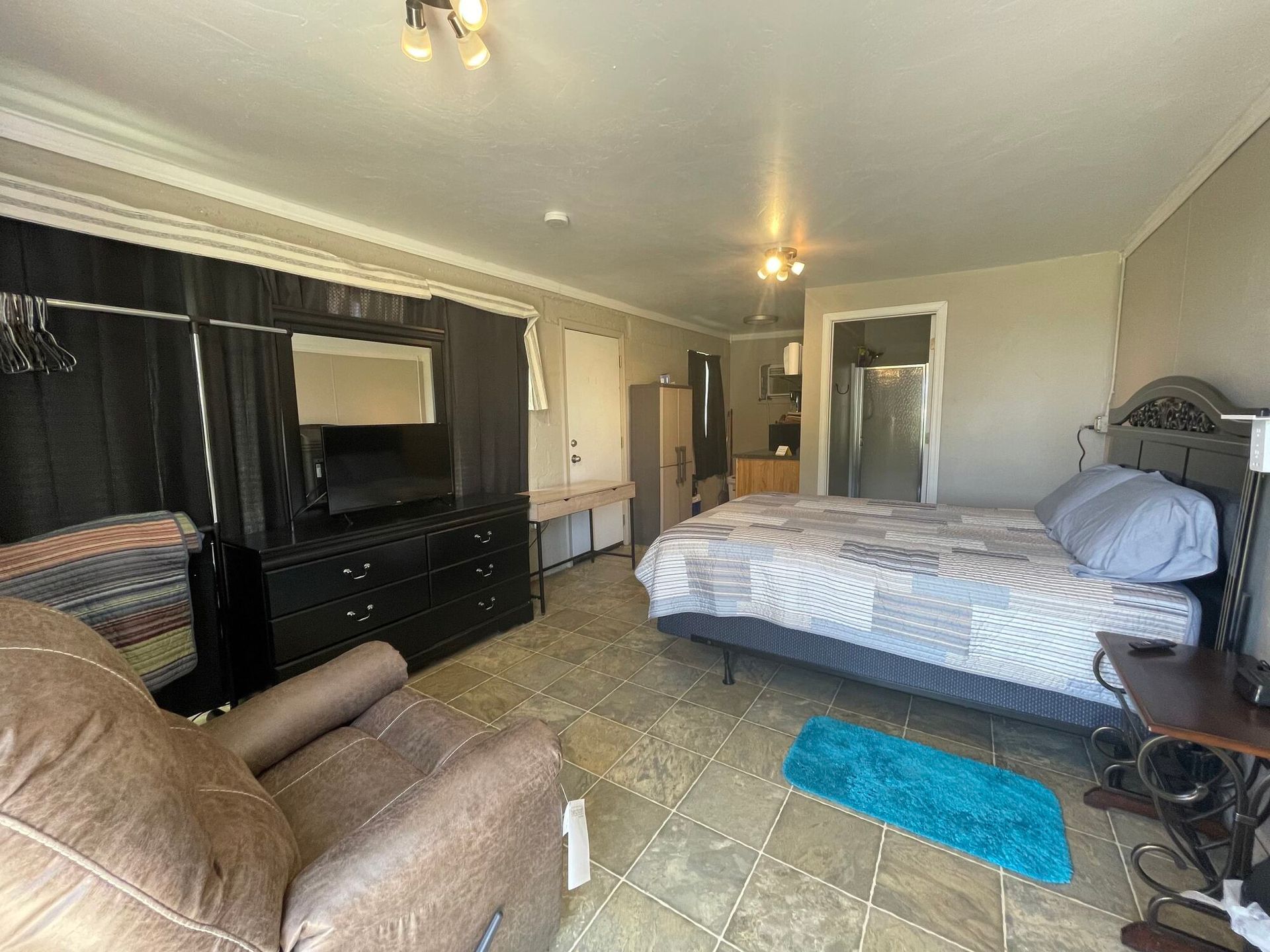 Spacious bedroom with bed, dresser, armchair, and blue rug under ceiling lights.