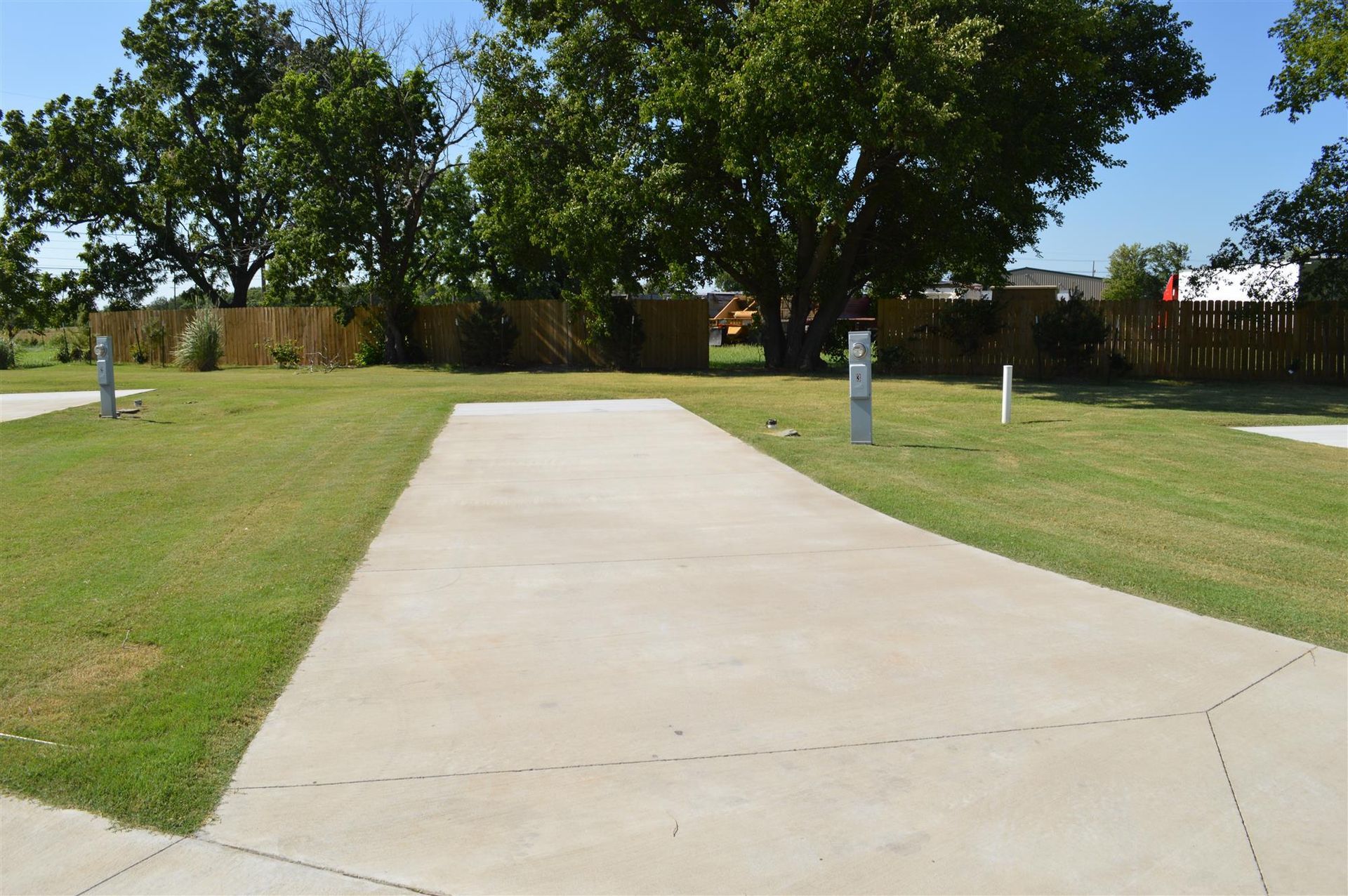 Concrete sidewalk path leading through a grassy park toward trees and a fence under a clear sky