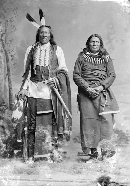 Two Native American people in traditional clothing stand side by side in a studio portrait.