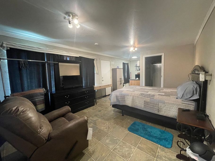 Bedroom with a bed, recliner, dresser, TV, and blue rug in a dimly lit room.