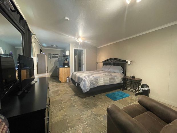 Bedroom with a bed, dresser, TV stand, and chair in a beige room with tiled floor.