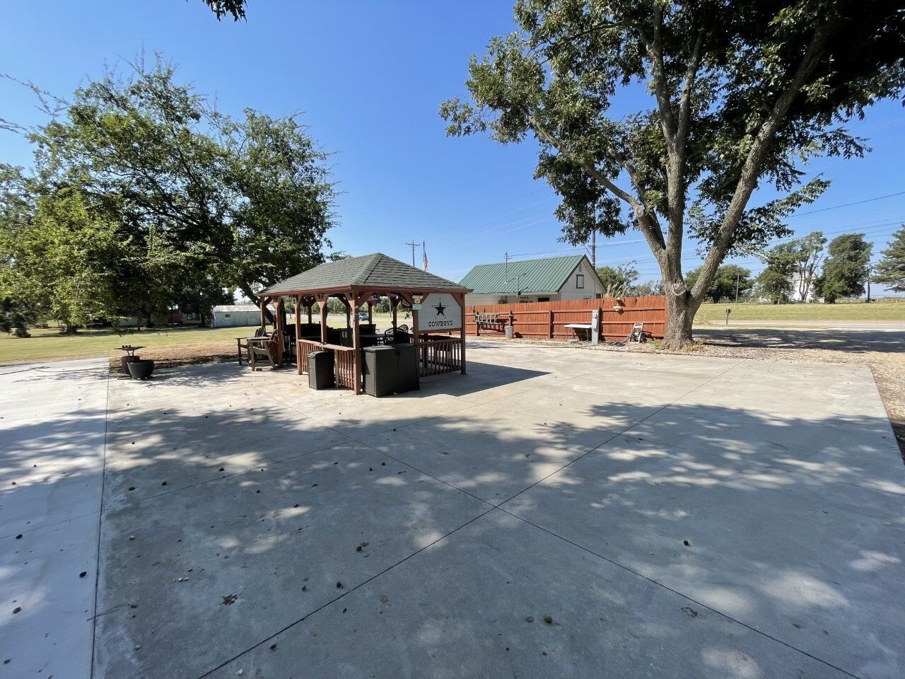 Shaded picnic pavilion with tables on a sunny concrete pad, surrounded by trees and open grassy parkland.