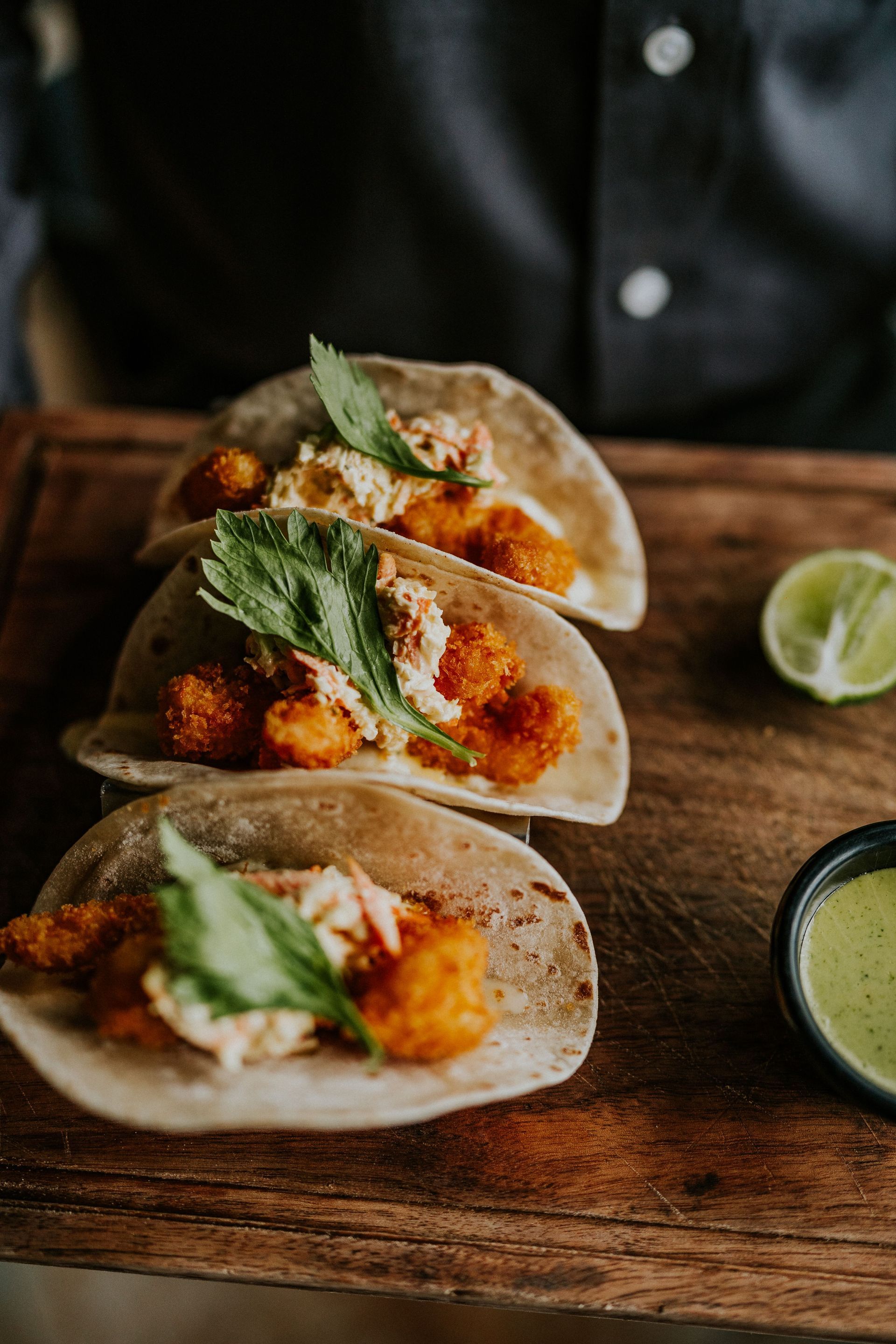 Three tacos with orange filling and cilantro on a wooden board, with lime and green sauce.