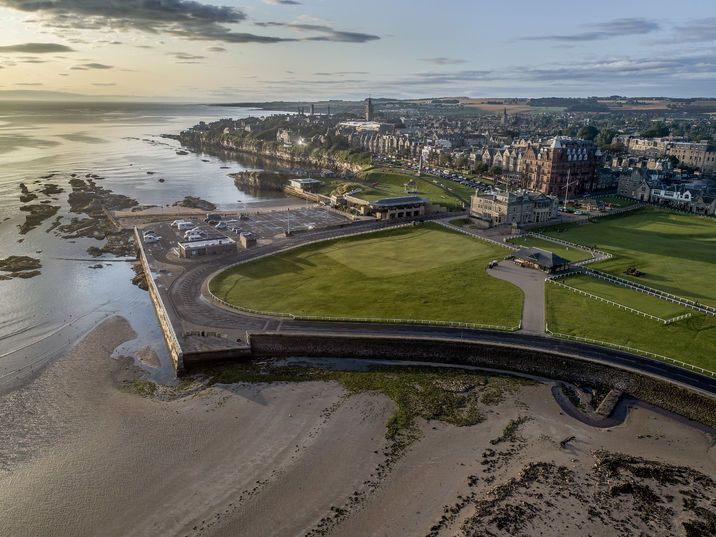 Aerial view of a coastal town with a sandy beach, seawall, and green park at sunset.