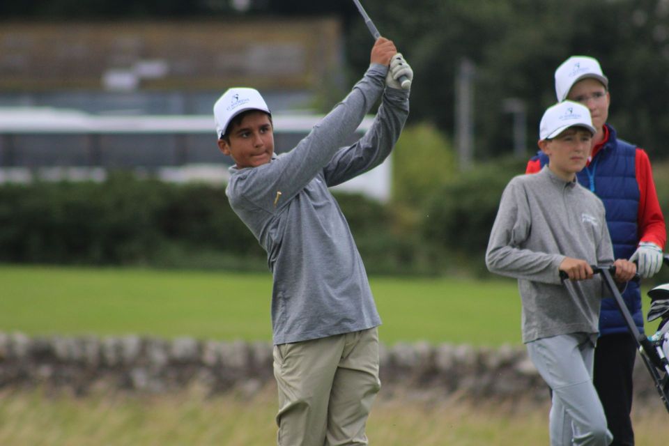 Golfer in gray shirt and cap swings club on a course, with two others watching nearby.