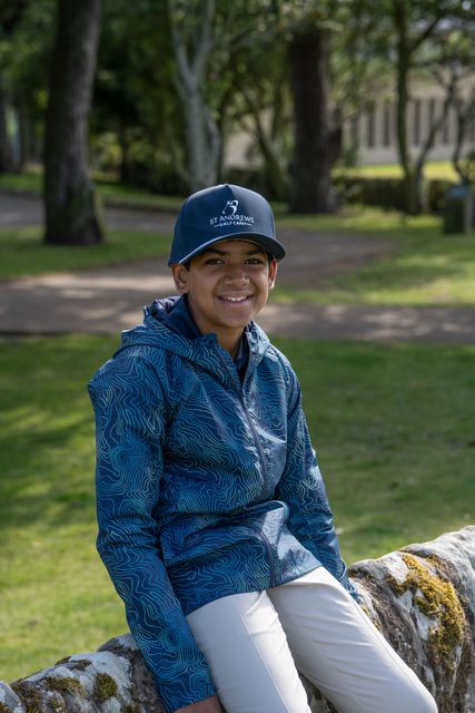 Person in a blue shirt and cap sitting on a mossy wall in a park, smiling.