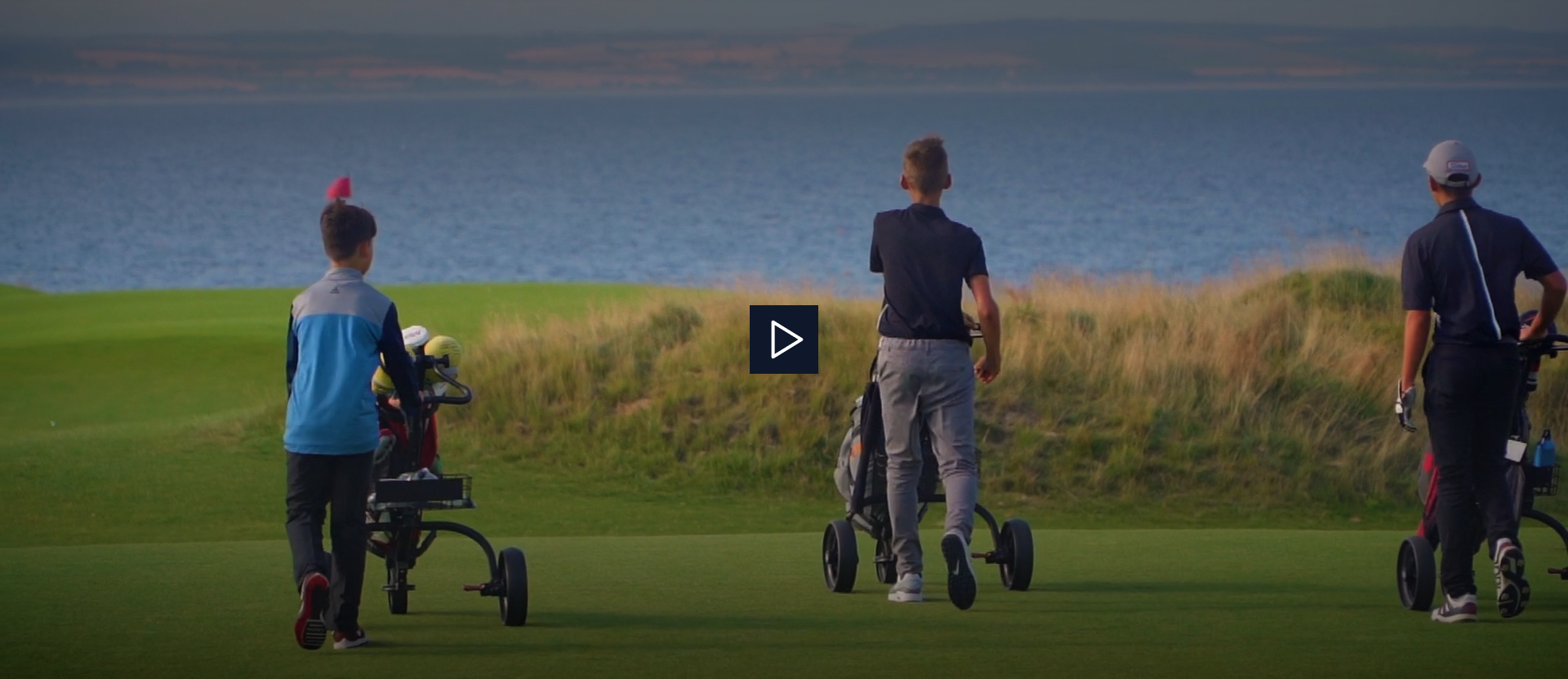 Three golfers standing on a green facing the ocean, with clubs and a golf cart nearby.