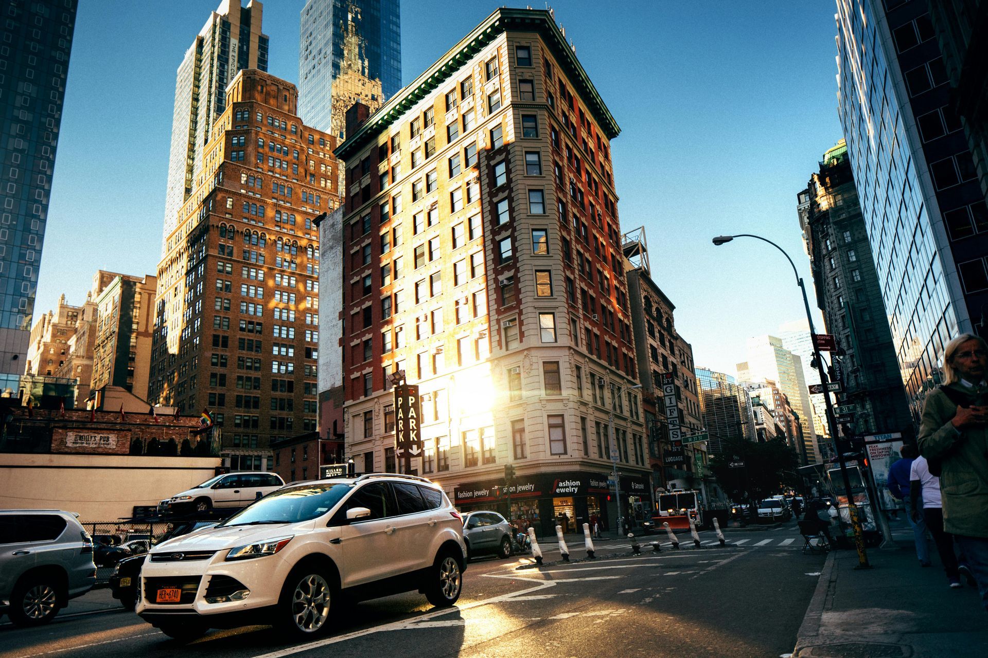 Sunlit city street with cars, tall buildings, and pedestrians in an urban intersection