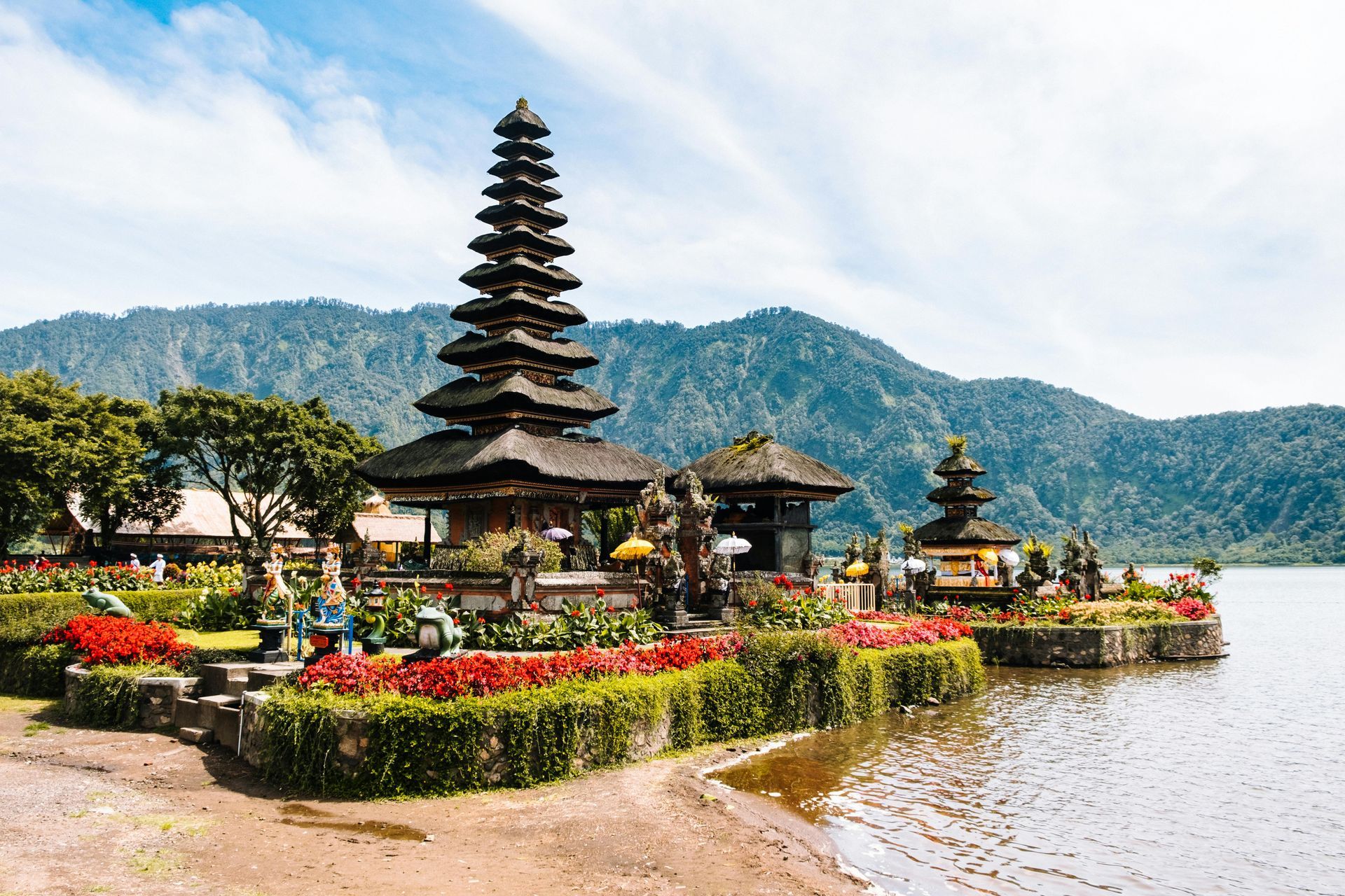 Balinese temple by a lakeshore with a tiered pagoda, flowers, and mountains in the background