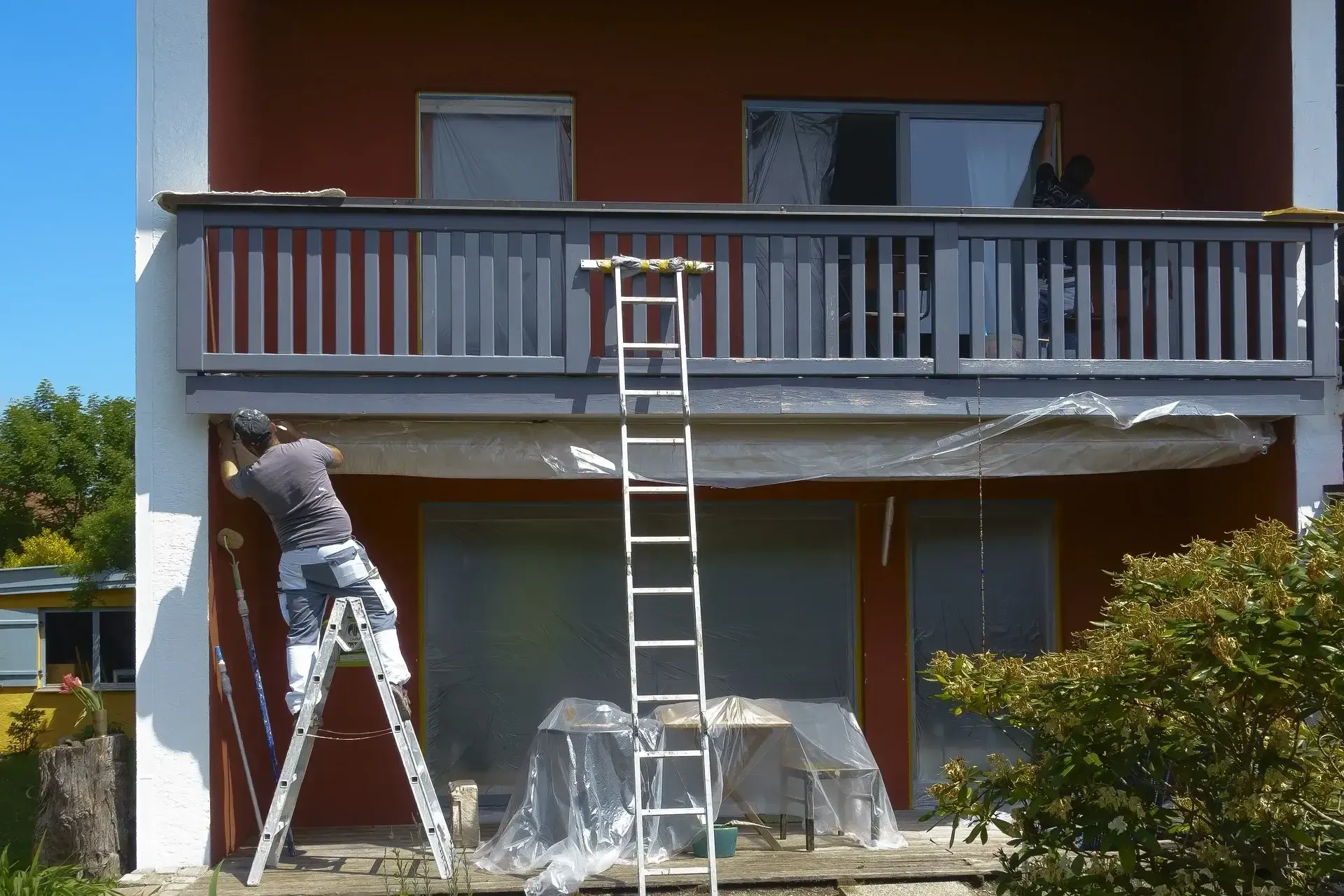 A man on a ladder is painting a balcony of a house.