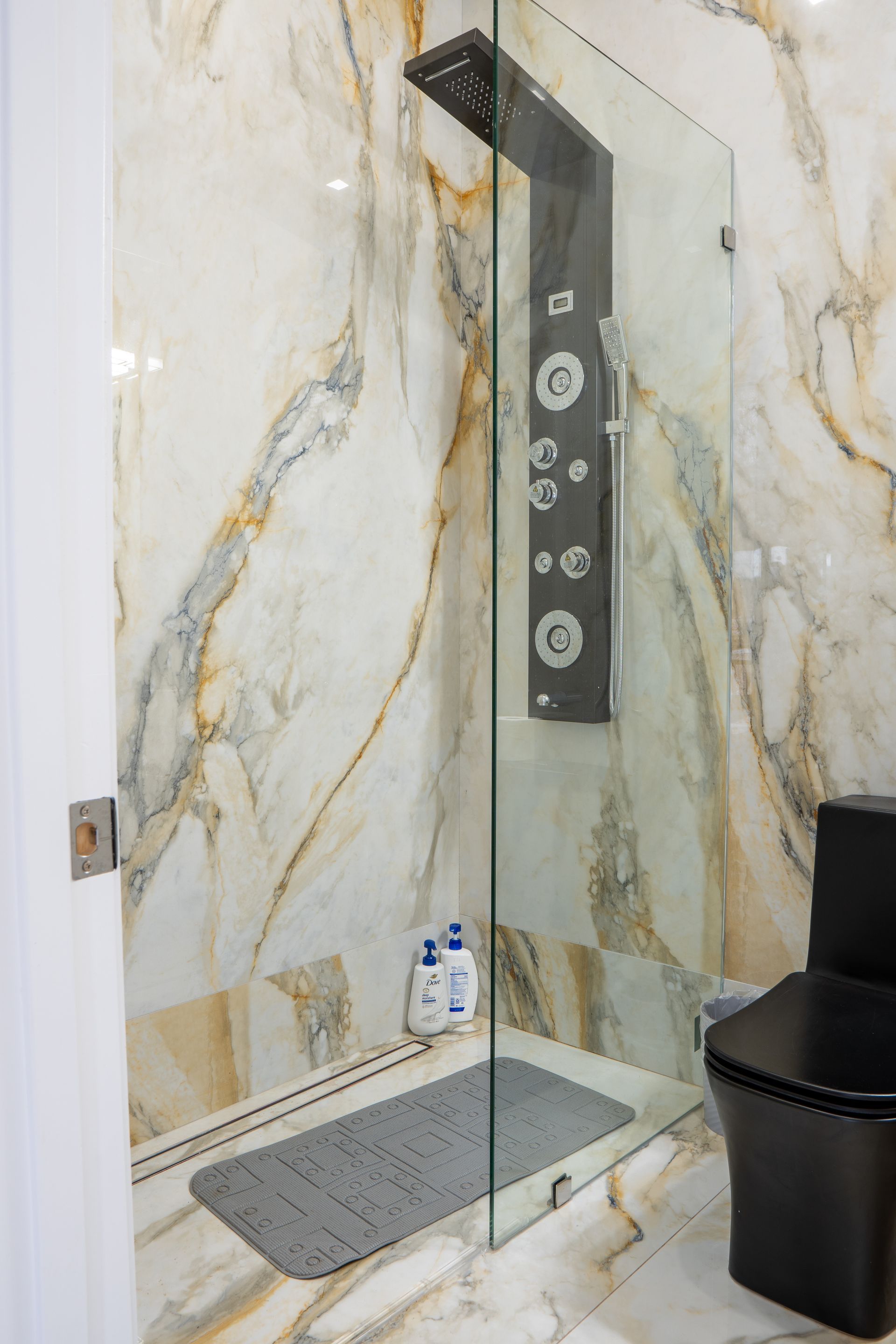 Shower with marble walls, glass door, and black showerhead; gray mat and black toilet visible.