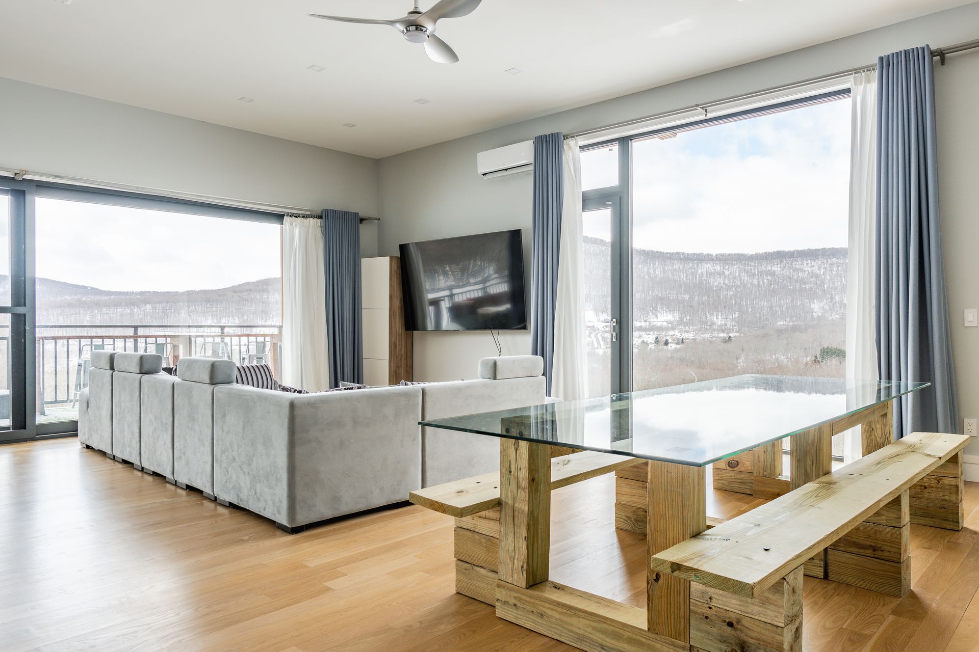 Living room with glass-top table, light-colored sectional sofa, large windows with mountain view, and air conditioner.