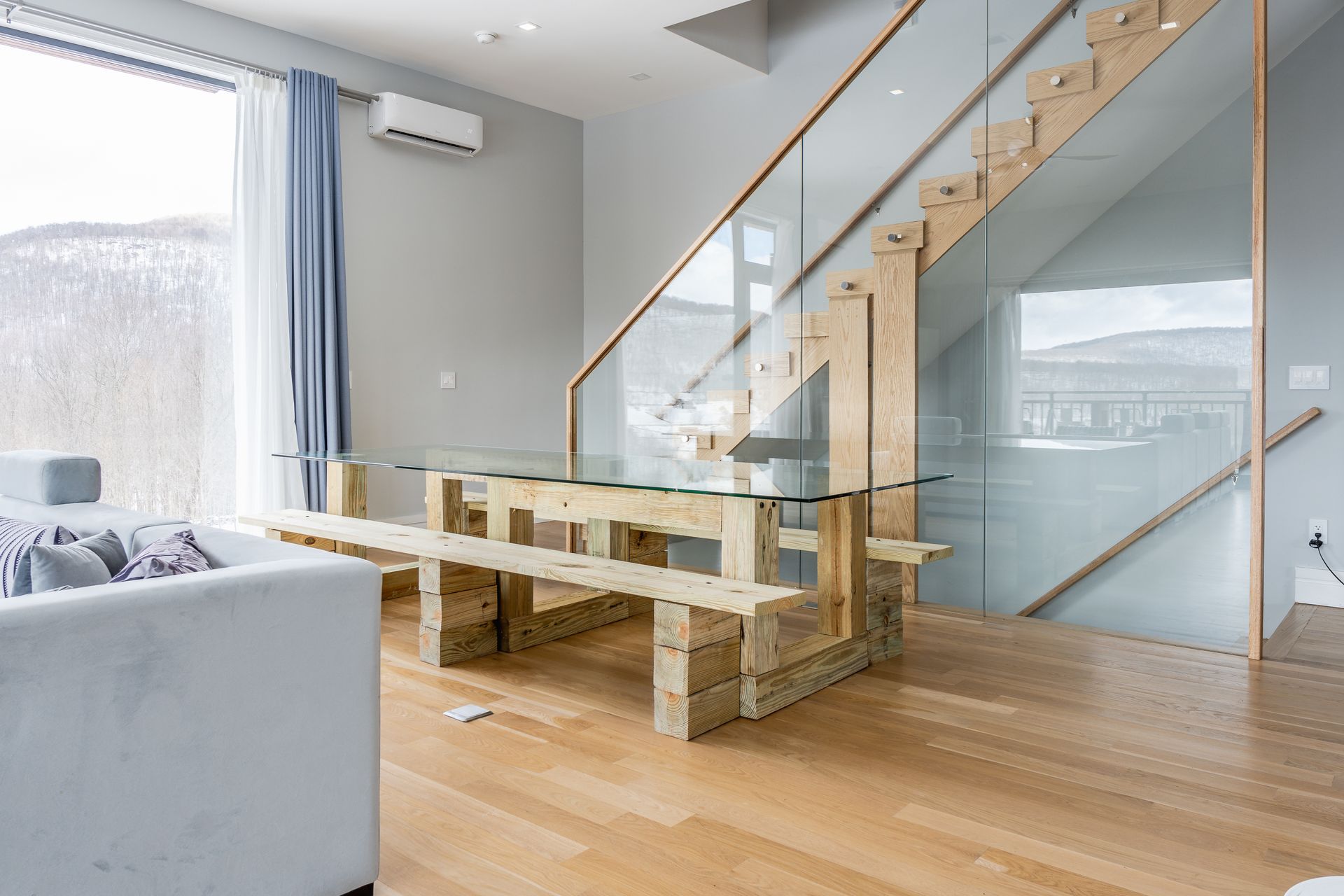 Dining area with glass tabletop, wooden benches, and a floating staircase. Large window with a mountain view.