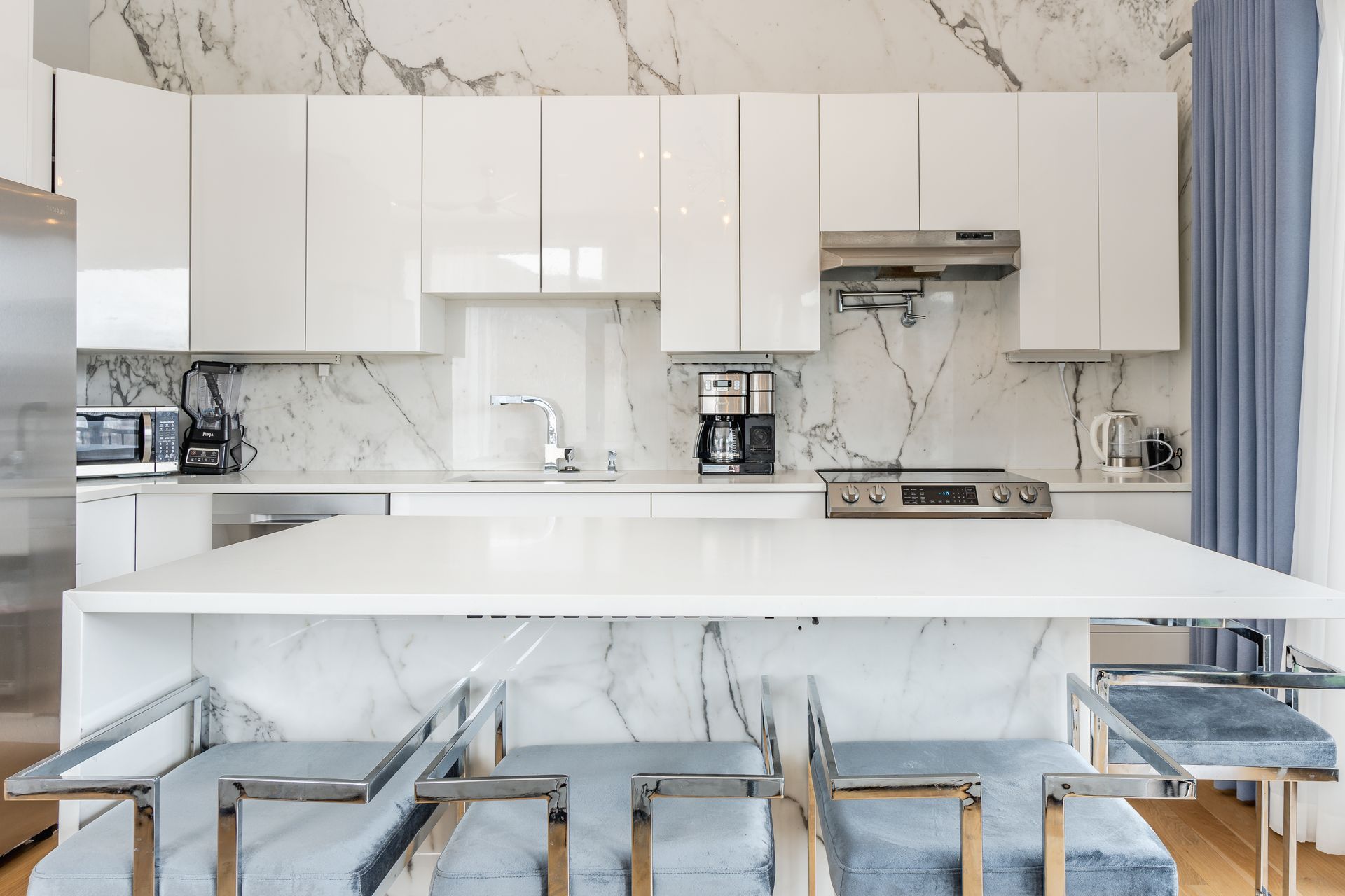 Modern white kitchen with marble accents and island, stainless steel appliances.