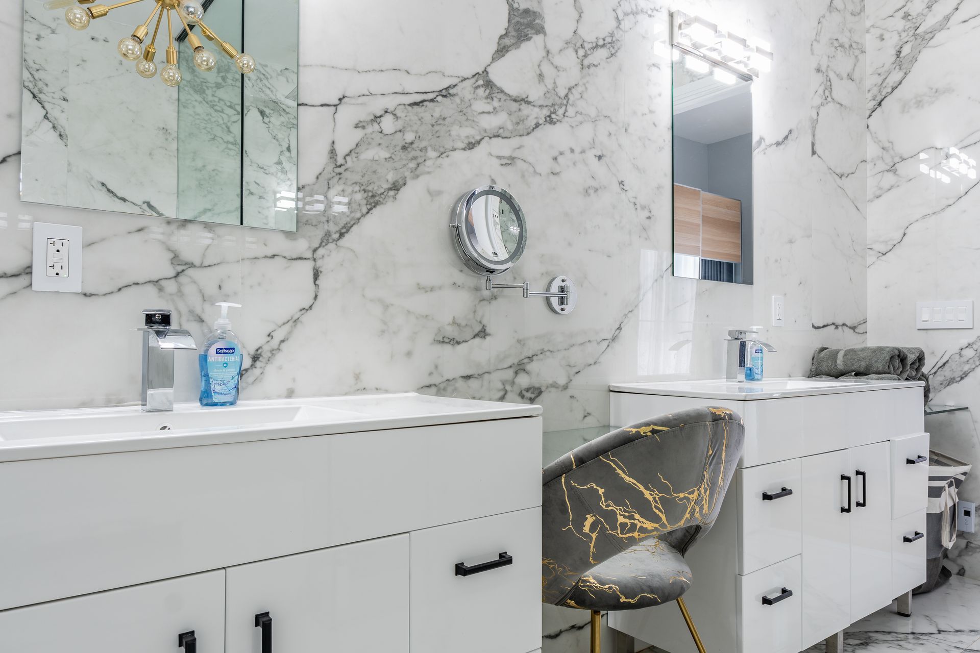 White bathroom with marble walls and countertops, two sinks, and a decorative chair.