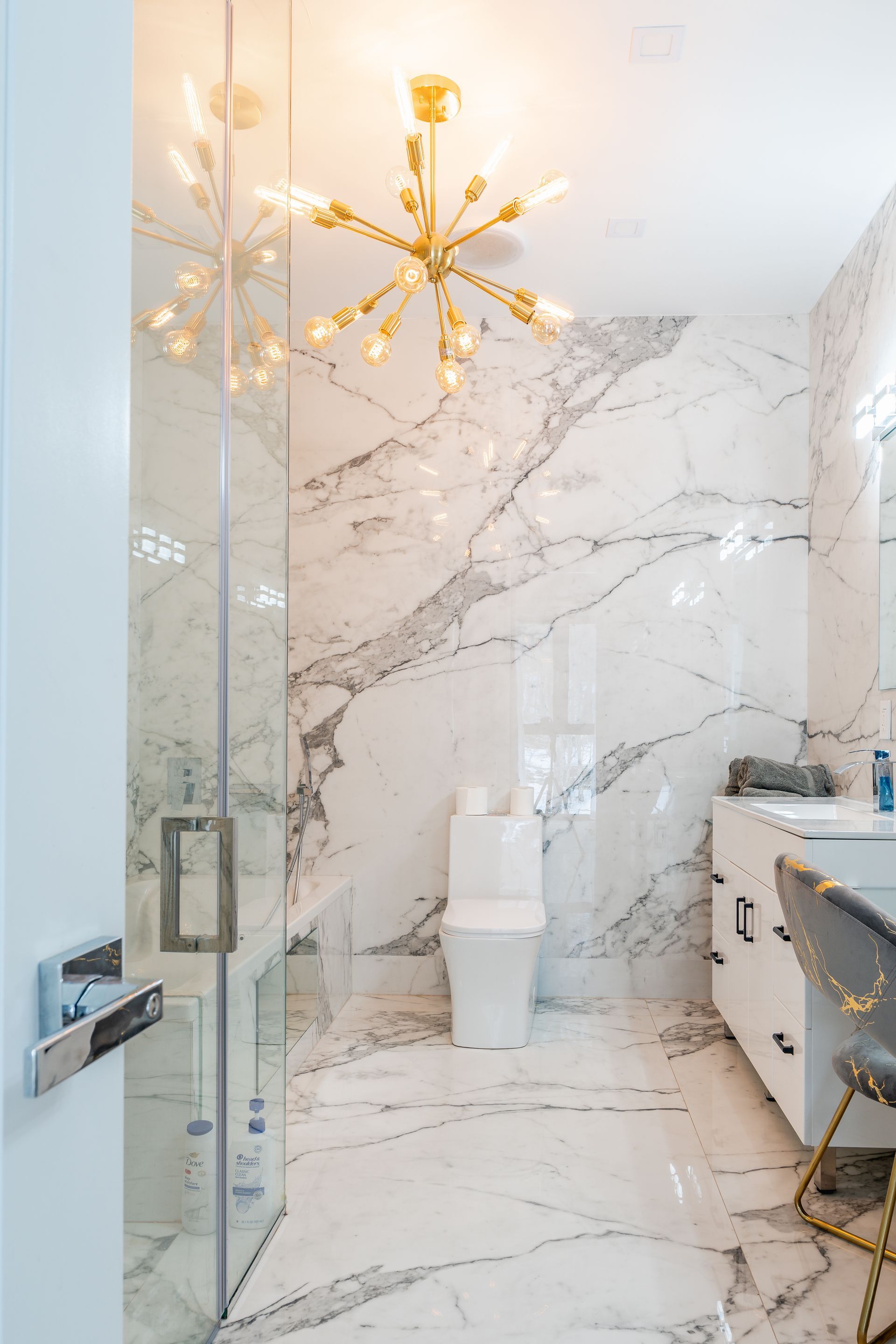 Luxurious bathroom with marble walls and floor, glass shower, and gold chandelier.