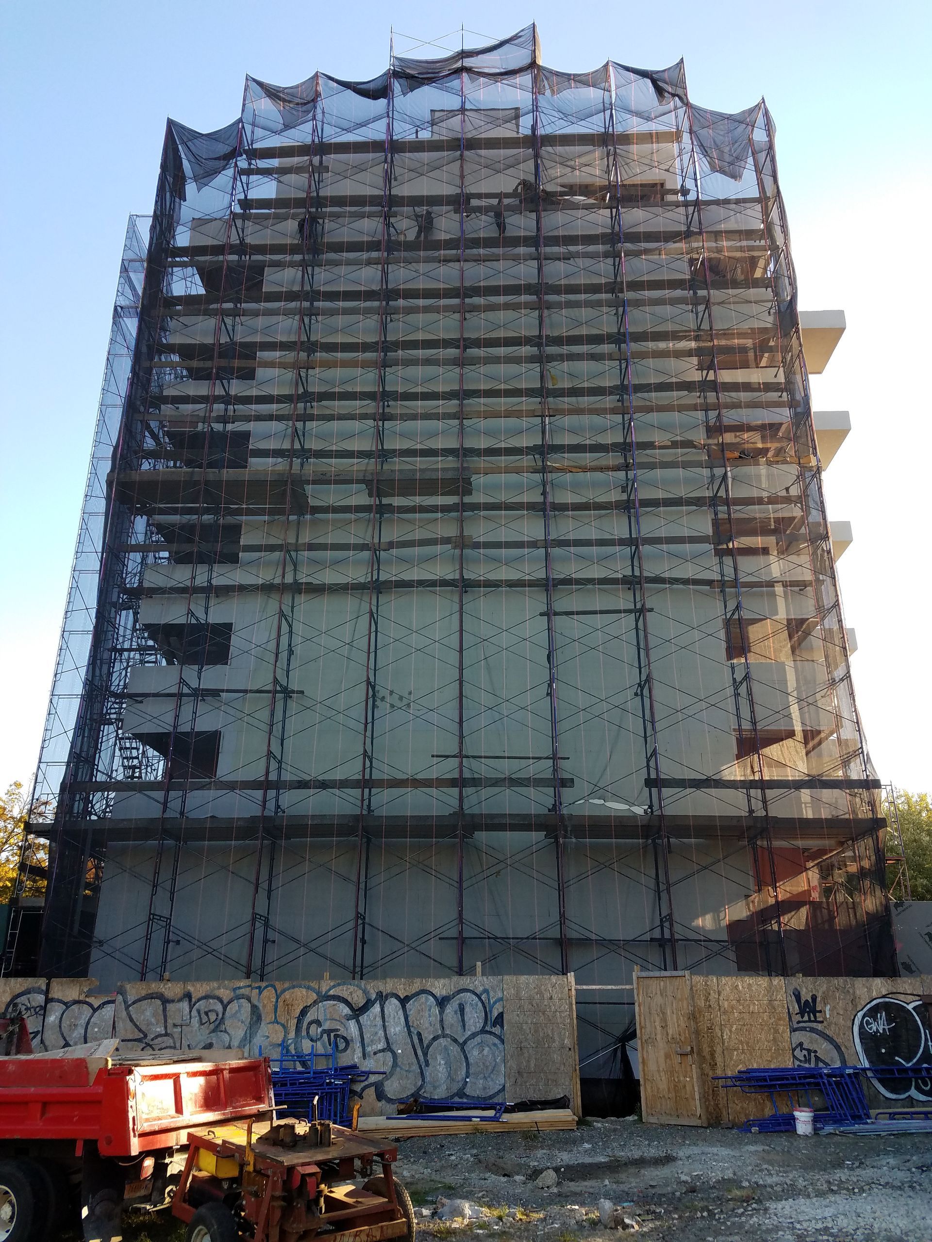 Building under construction, covered in scaffolding and black netting. Construction materials are at the base.