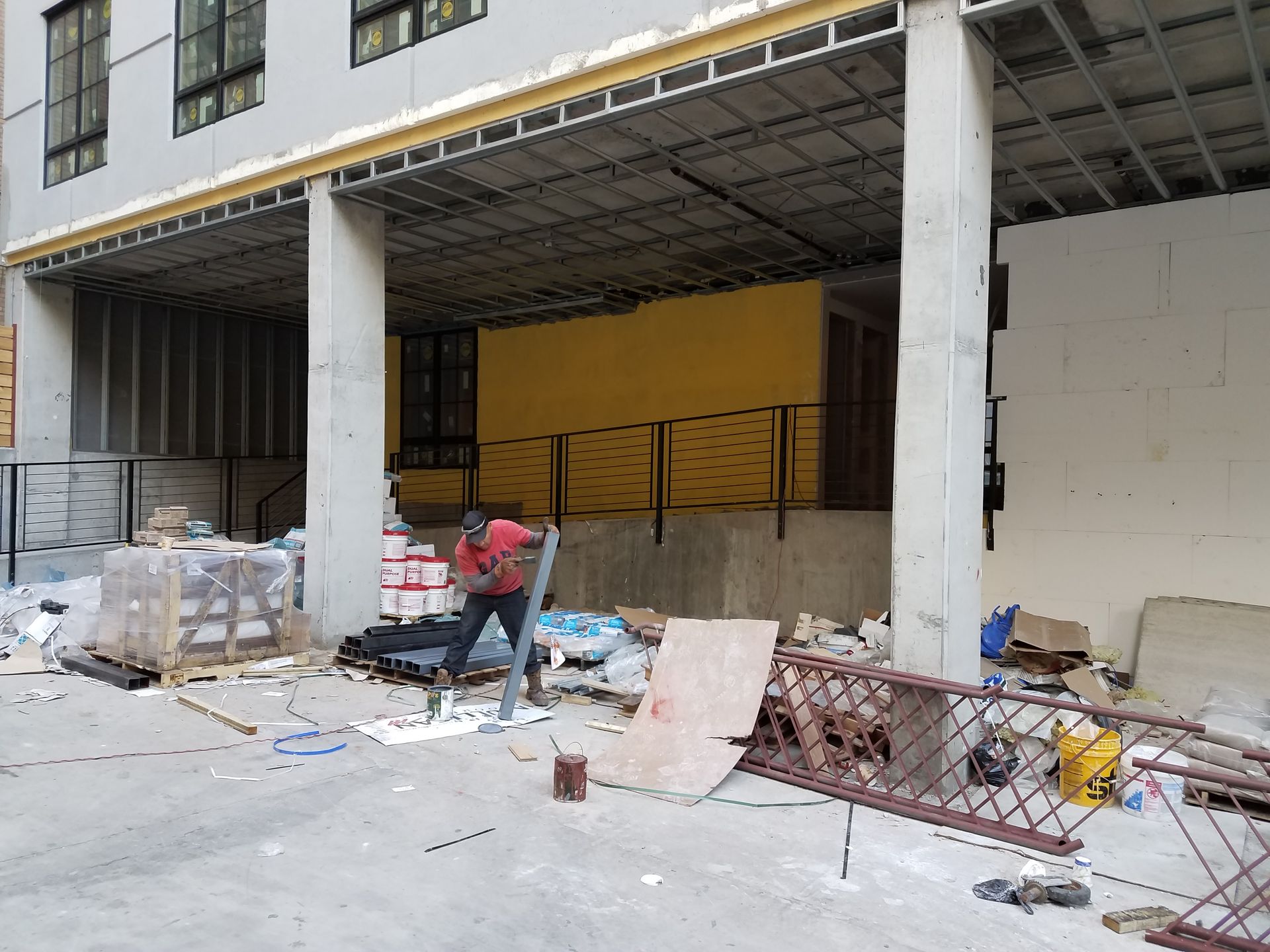 Construction site with a person working. Under a building with gray pillars is a yellow wall and a ramp. Debris litters the ground.