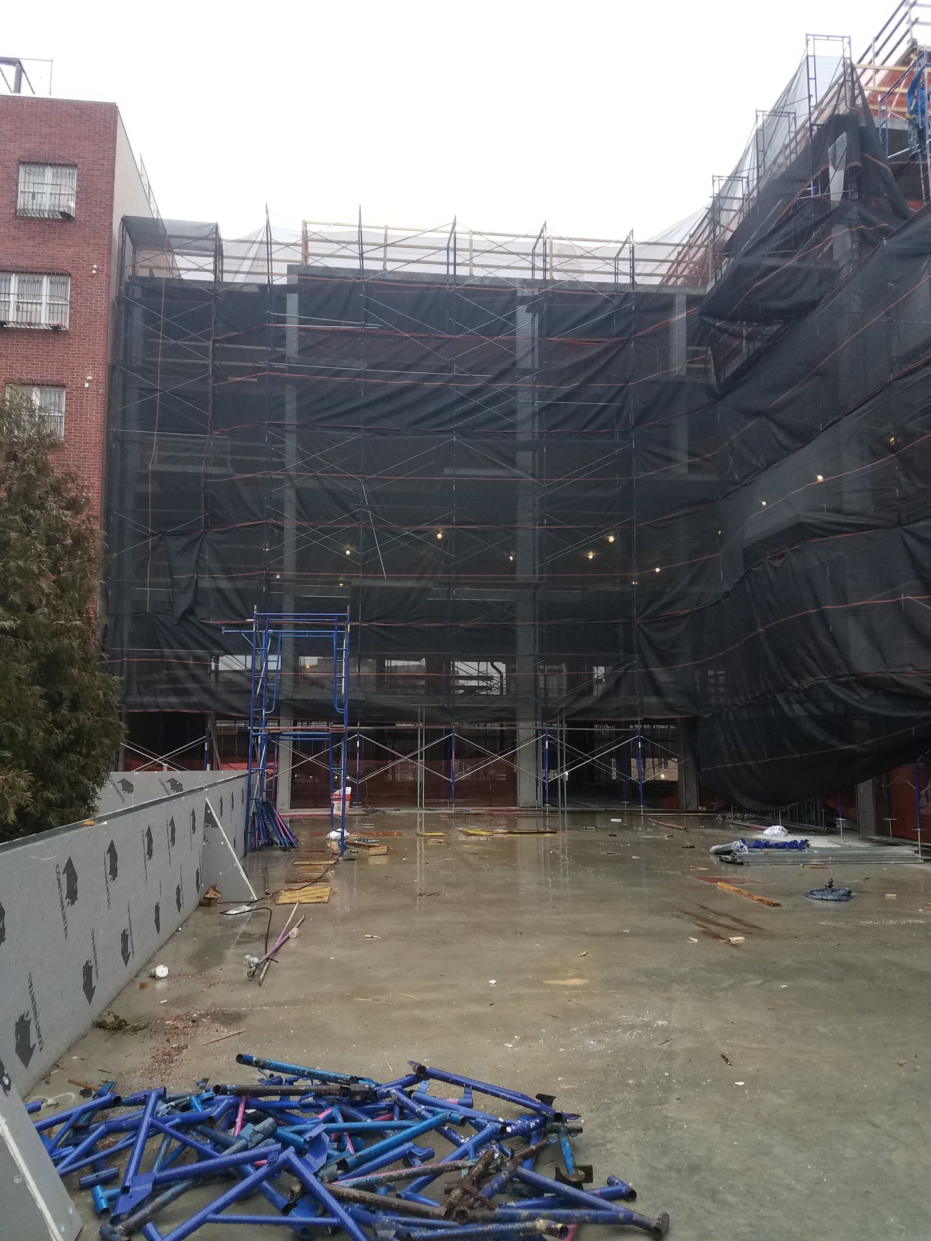 Construction site with a partially built building covered in black netting and scaffolding.