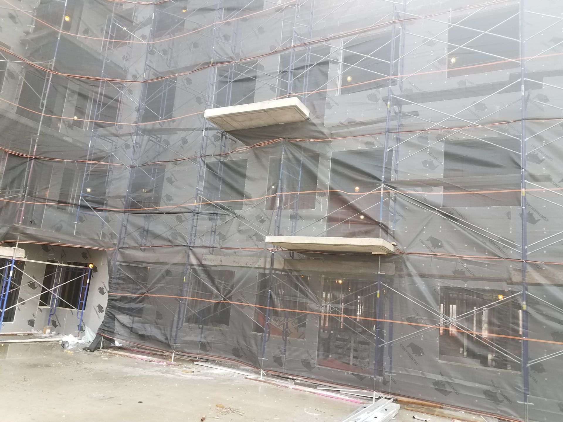 Building exterior under construction, shrouded in black netting and scaffolding. Concrete platforms extend.