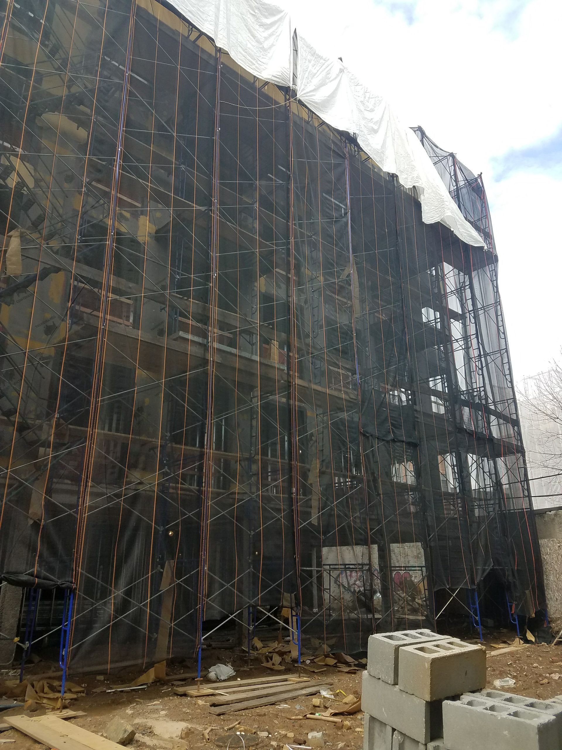 Building under construction, covered in black netting and white tarp.  Brick blocks in foreground.
