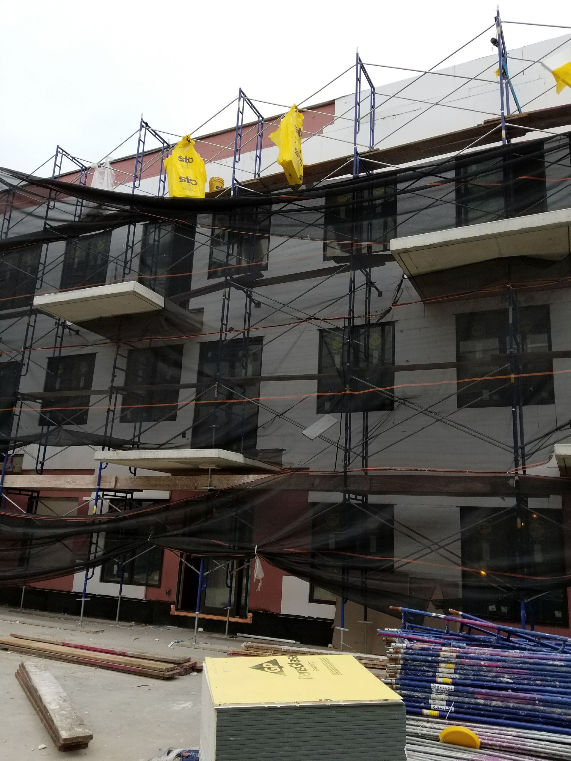 Building under construction, scaffolding with safety netting; gray and white exterior; yellow safety markers.