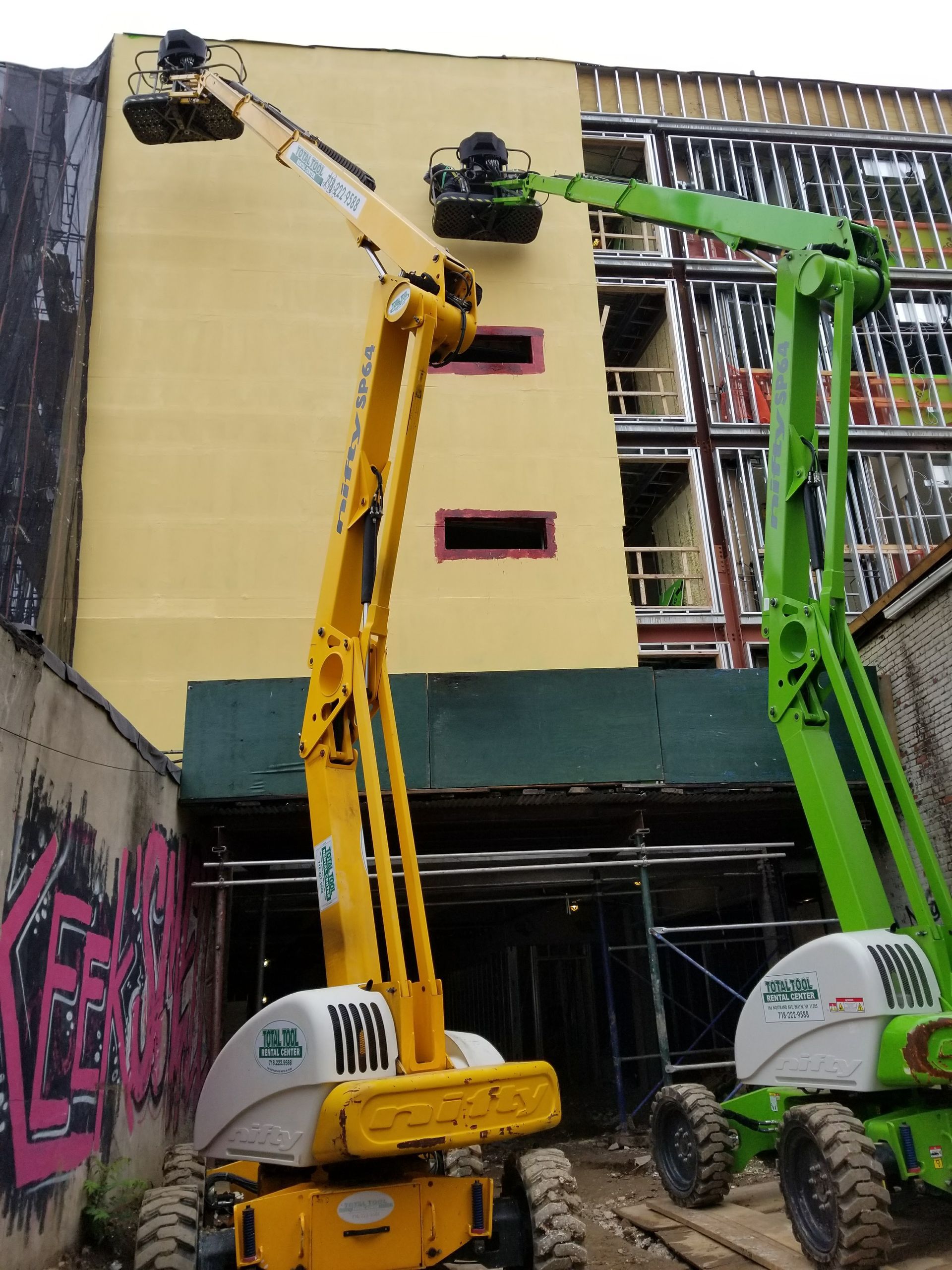 Yellow and green boom lifts near a building with construction, graffiti on the left wall.