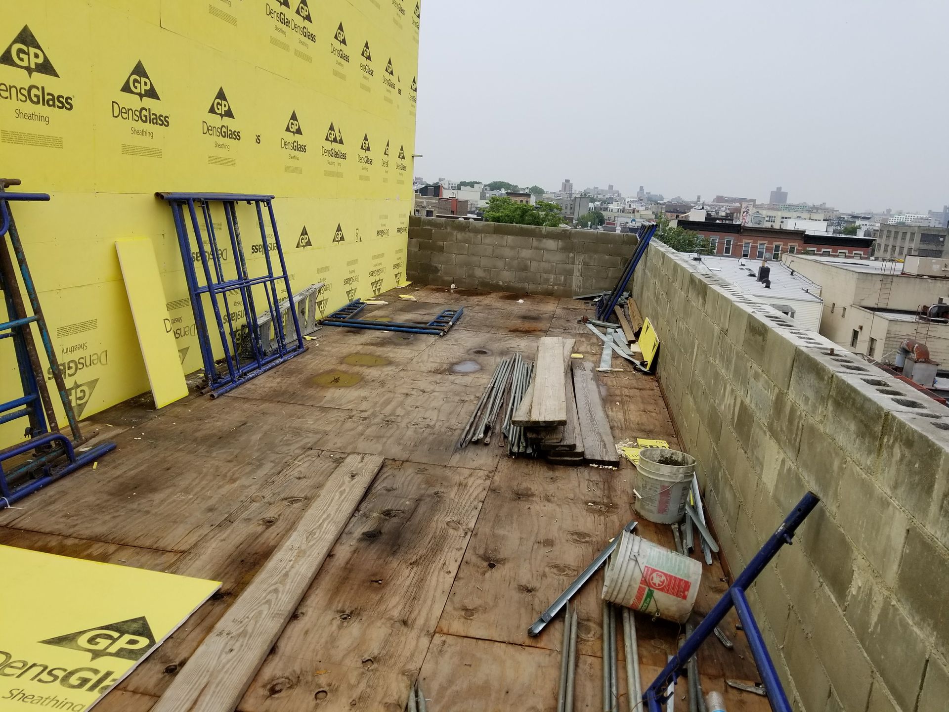 Construction site on a rooftop; yellow sheathing, scaffolding, wood planks, and concrete block walls. City in background.