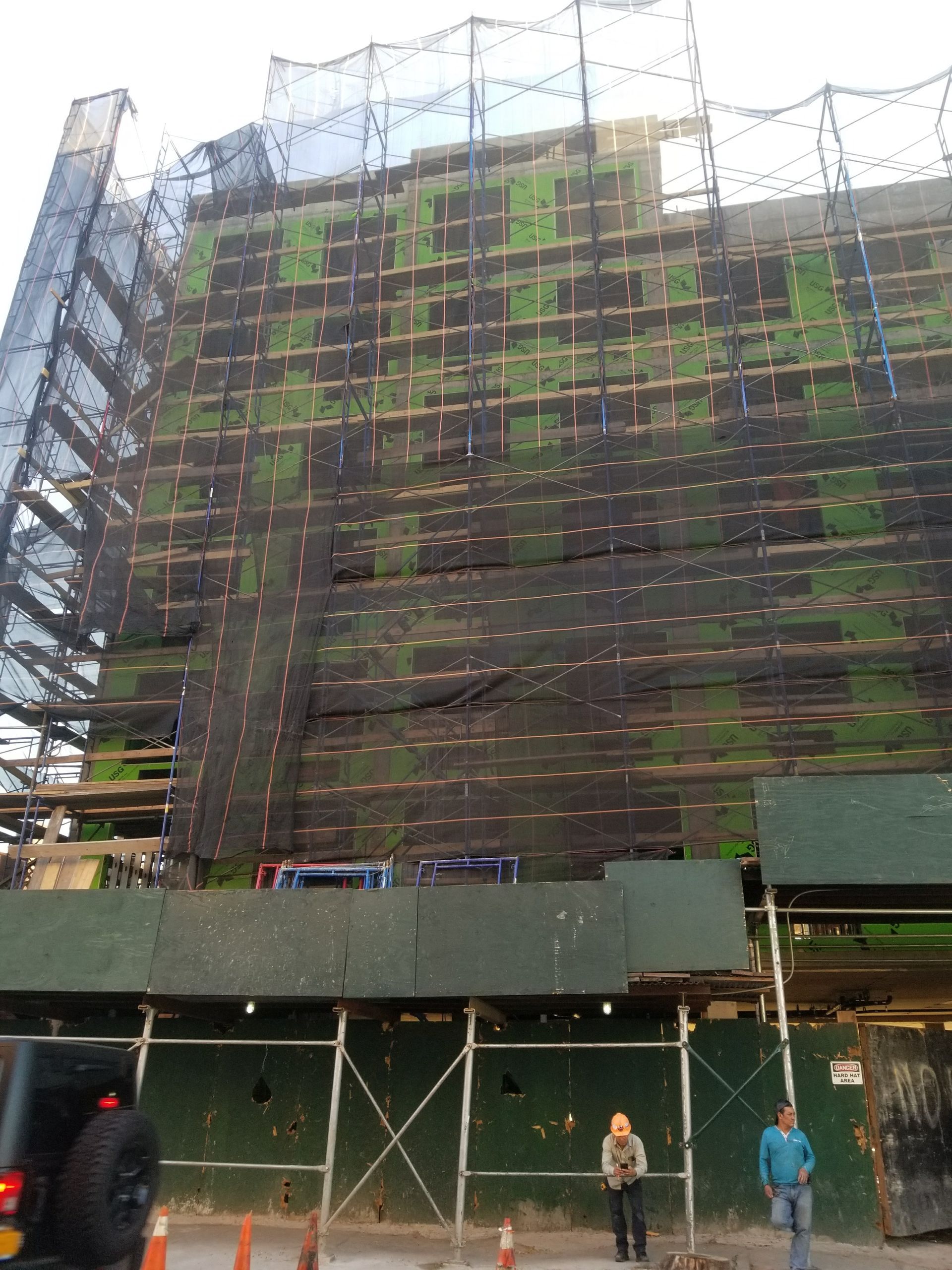 Building under construction, covered in scaffolding and safety netting; two people stand on the sidewalk.
