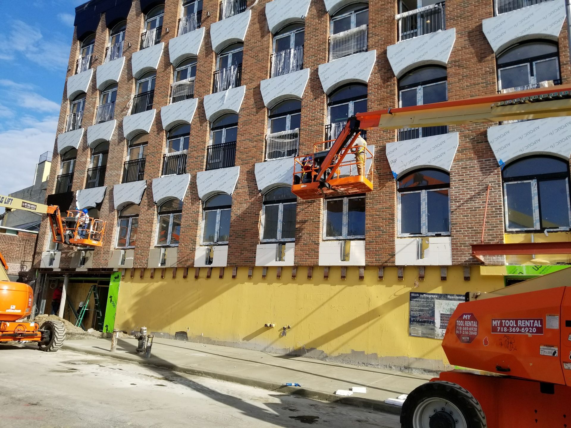 Building under construction; workers in orange lifts, brick facade with decorative window frames, yellow barrier.