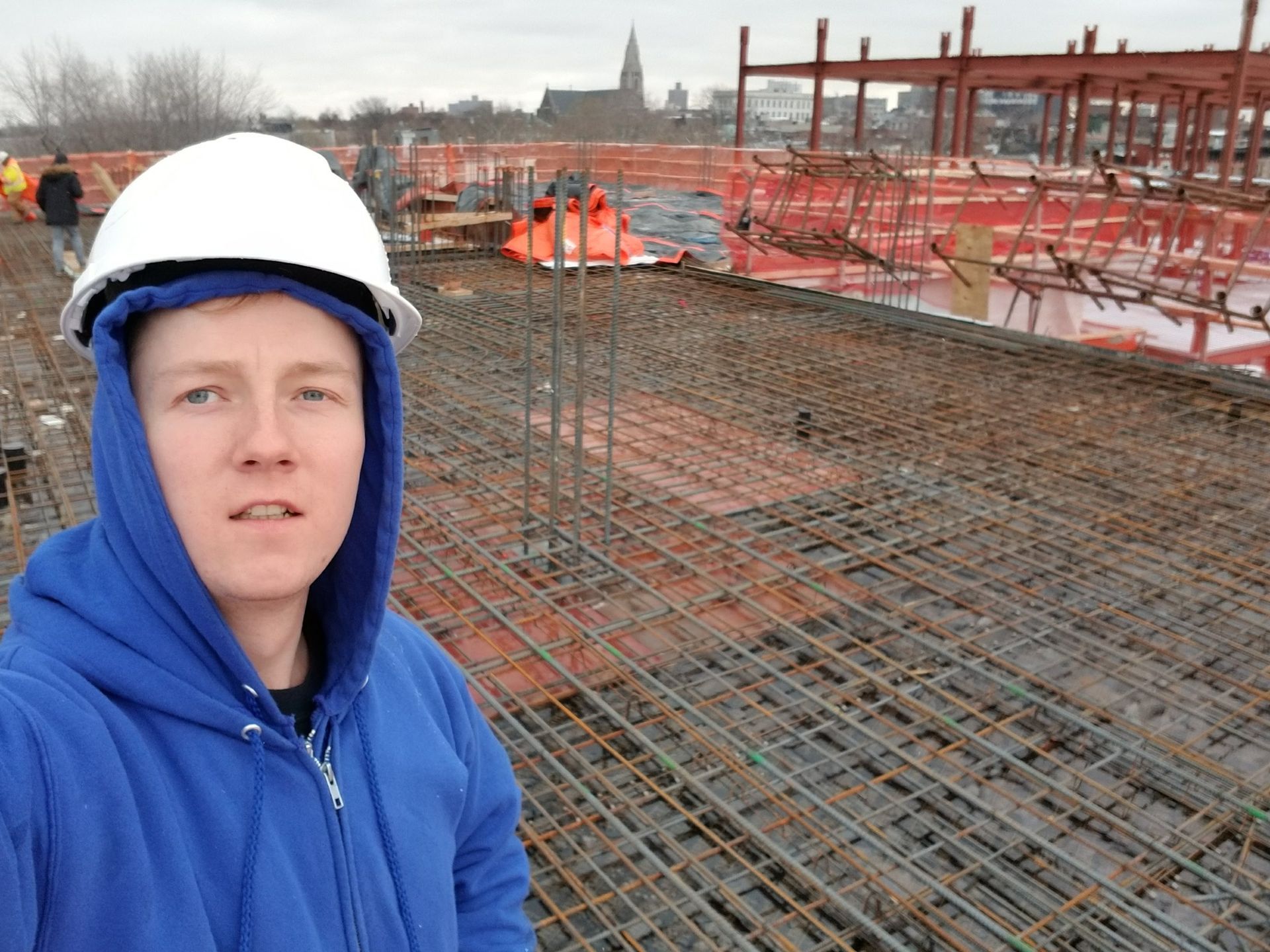 Construction worker in a blue hoodie and white hard hat on a rebar-filled construction site with orange and brown structures and a church in the background.
