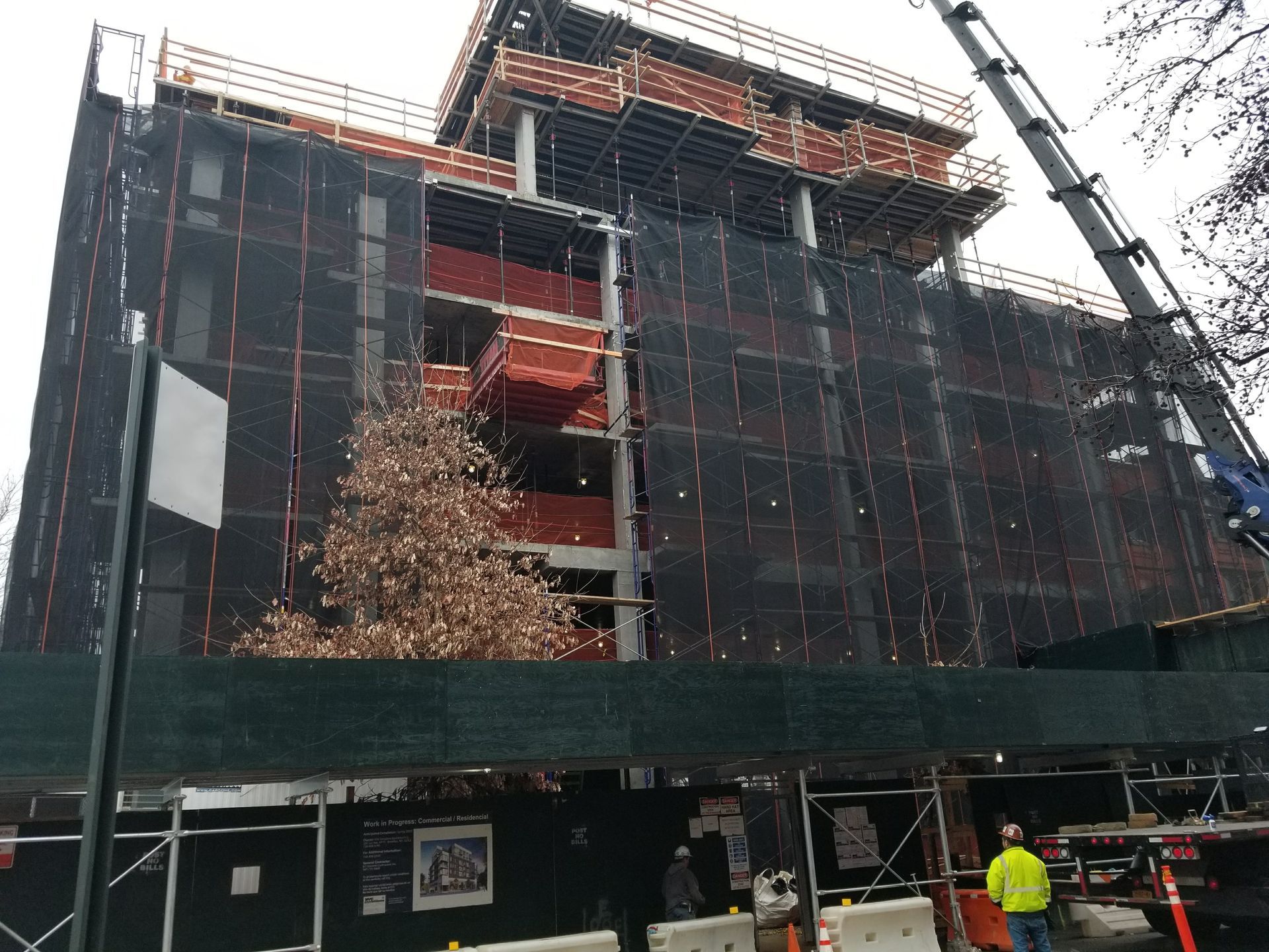 Building under construction, covered in black netting, with a crane on the right. A few workers are visible.
