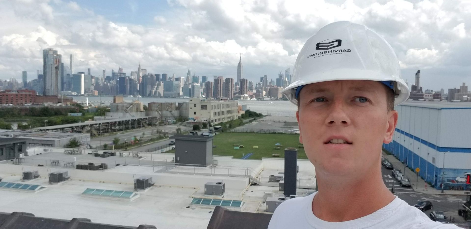 Man wearing a white hard hat, with a city skyline in the background on an overcast day.