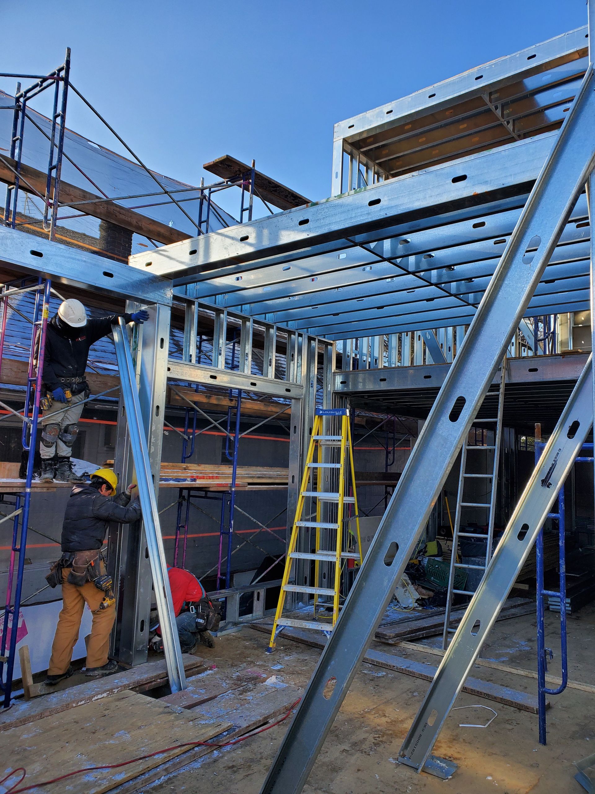 Construction site with metal framing. Workers in hard hats use tools. Scaffolding, ladders, blue sky.