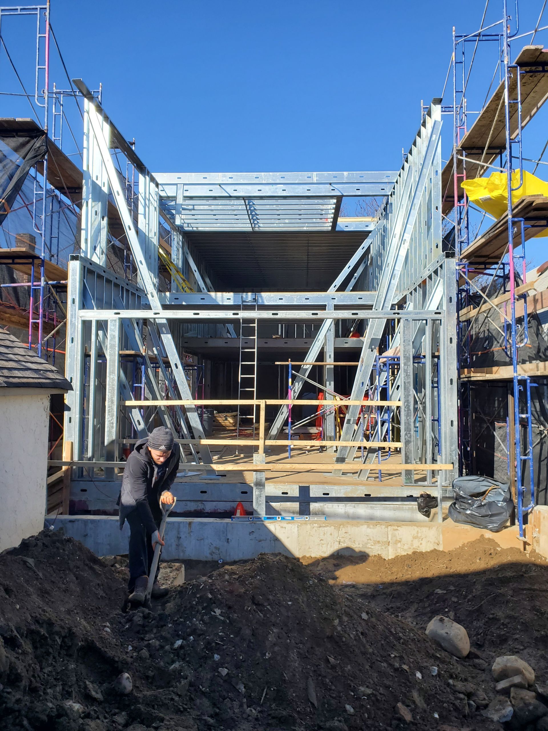 Construction site with steel framing. Worker digging in foreground. Scaffolding, blue sky.
